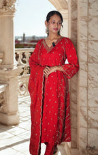 Woman wearing a red traditional outfit with intricate patterns, standing in a sunlit indoor setting.