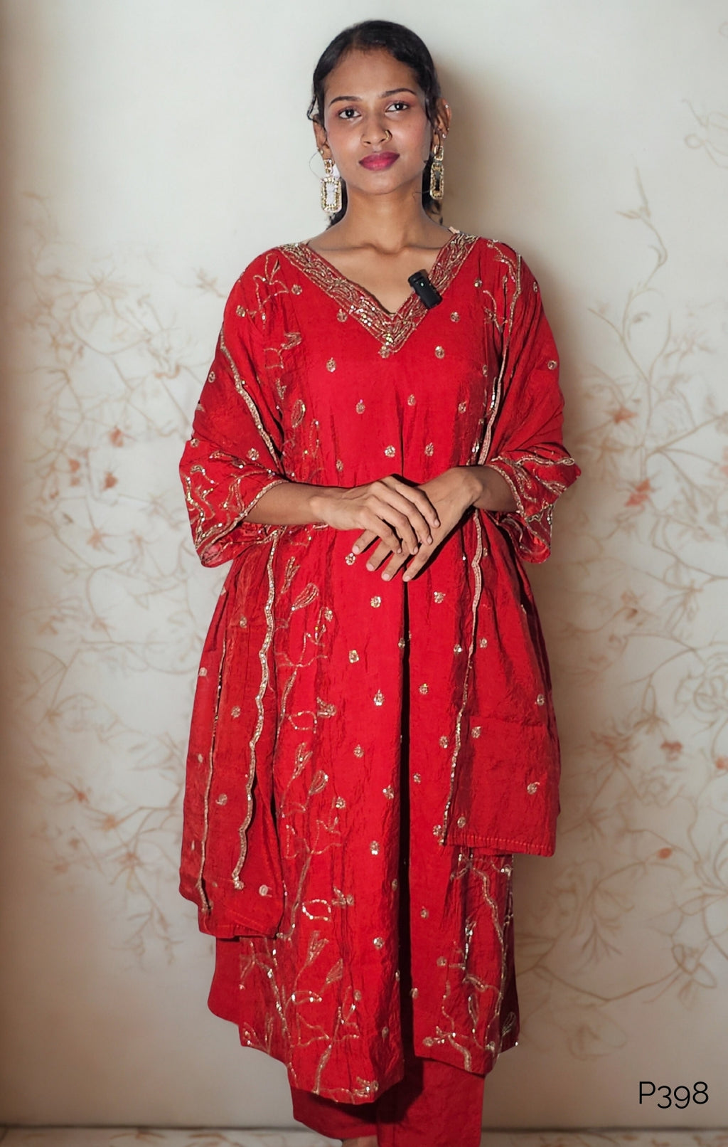 Woman wearing a red traditional outfit with white patterns against a light floral-patterned wall.