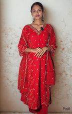 Woman wearing a red traditional outfit with white patterns against a light floral-patterned wall.