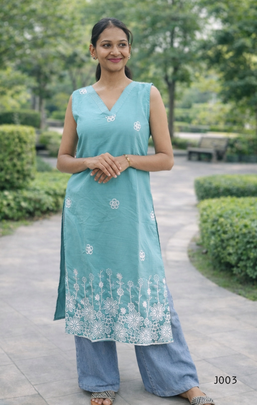 Woman wearing a teal sleeveless top and blue pants with white floral patterns, standing in a garden.