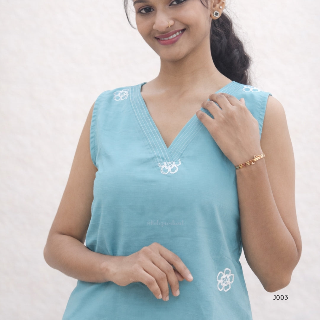 Woman wearing a light blue sleeveless top with white floral embroidery on a plain background