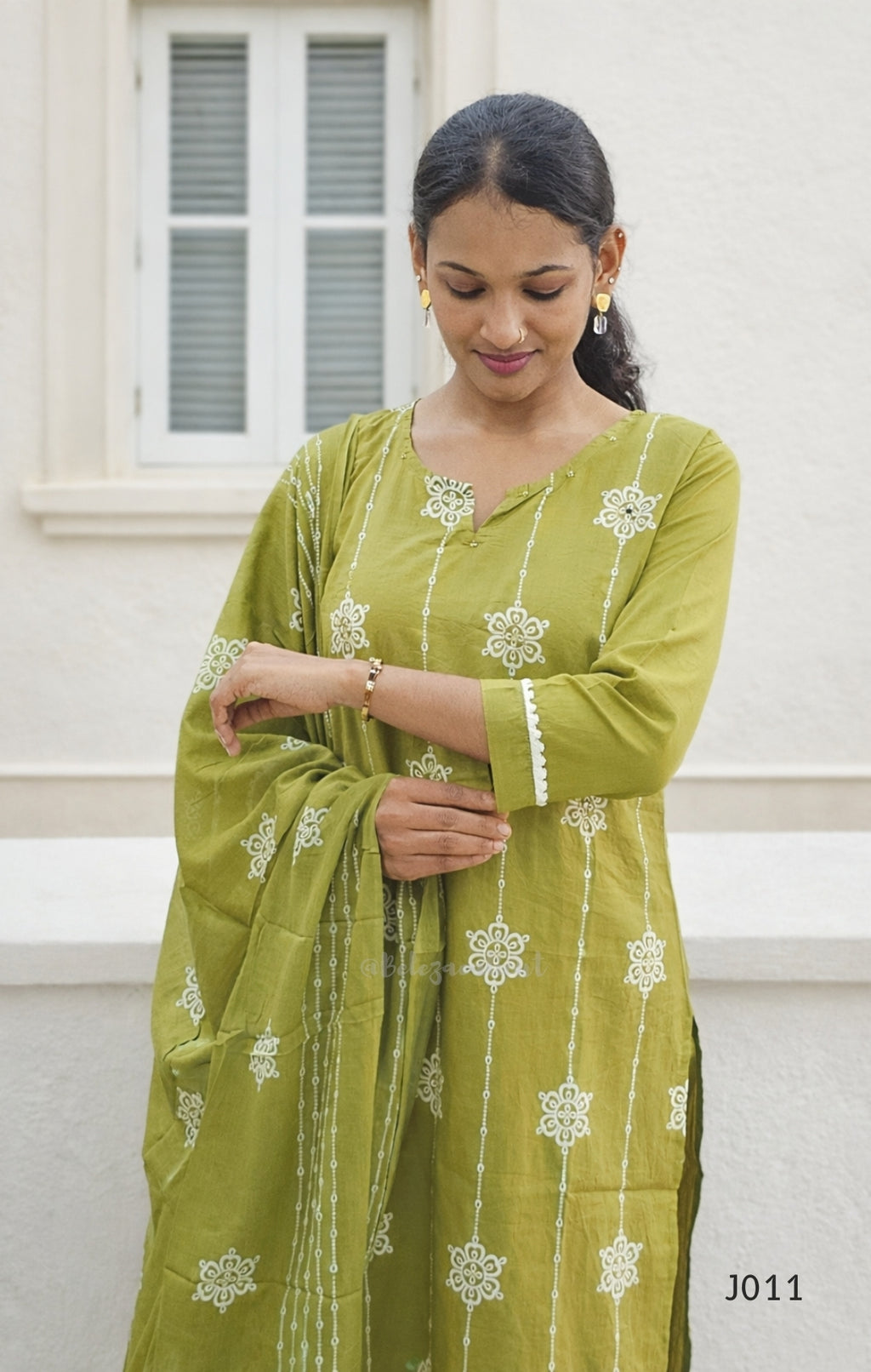 Woman wearing a green traditional outfit with white patterns against a white wall.