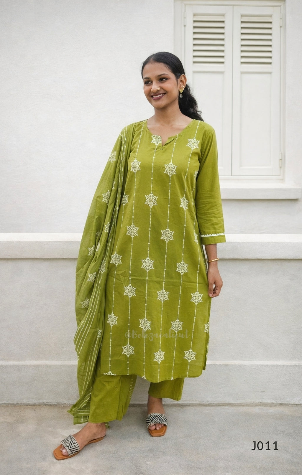 Woman wearing a green traditional outfit with white patterns against a white wall.