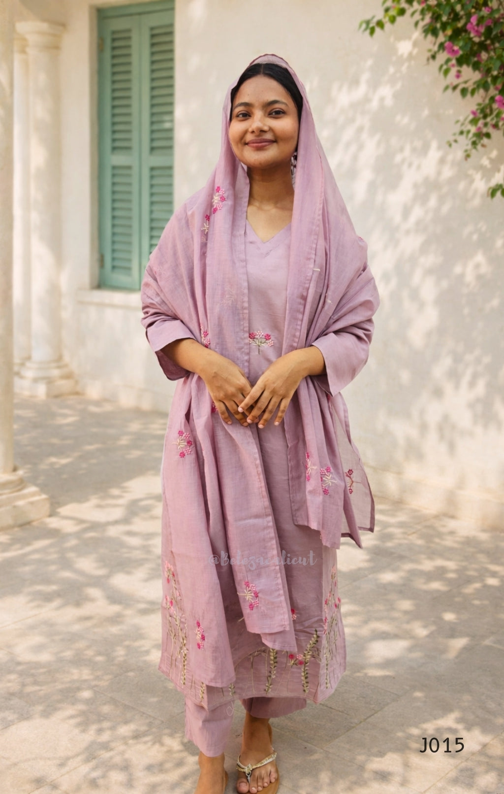 Woman in a pink traditional outfit standing outdoors with a building and greenery in the background.