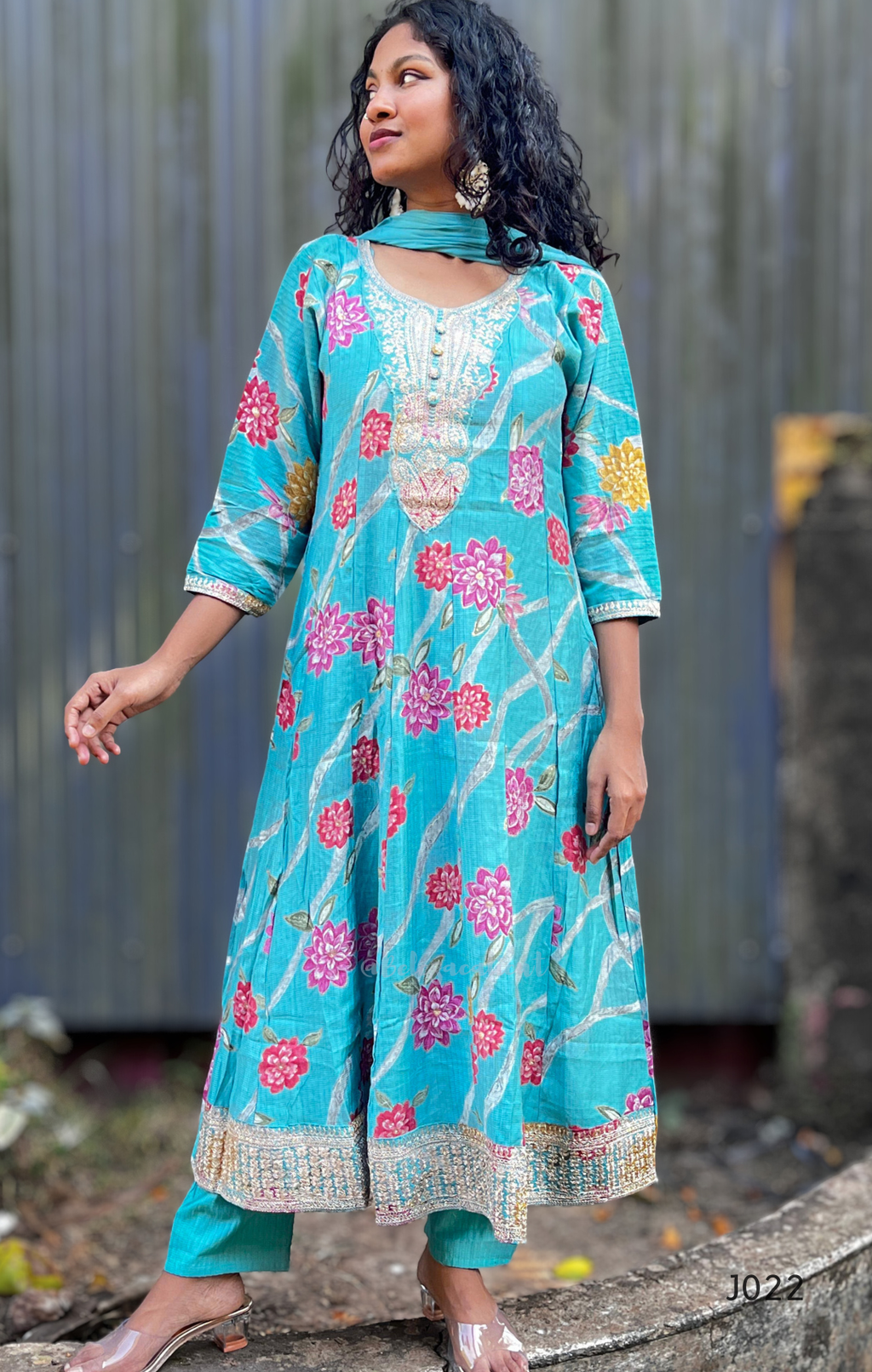 Woman wearing a turquoise floral dress standing against a metallic wall.