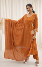 Woman wearing an orange traditional outfit with a matching dupatta against a white curtain background