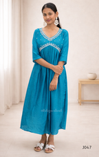 Woman wearing a blue dress with white embroidery in a minimal indoor setting.