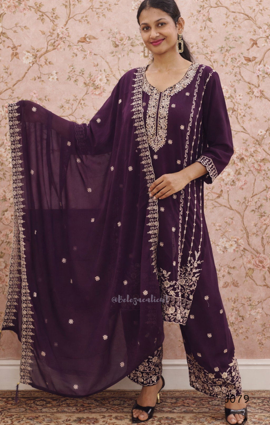 Woman wearing a purple traditional outfit with intricate designs against a floral wallpaper background