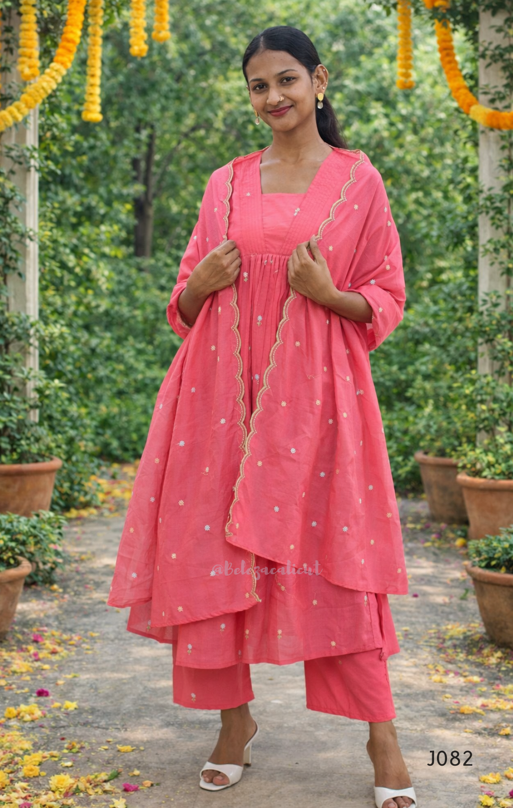 Woman in a pink traditional outfit standing outdoors with greenery and decorations.