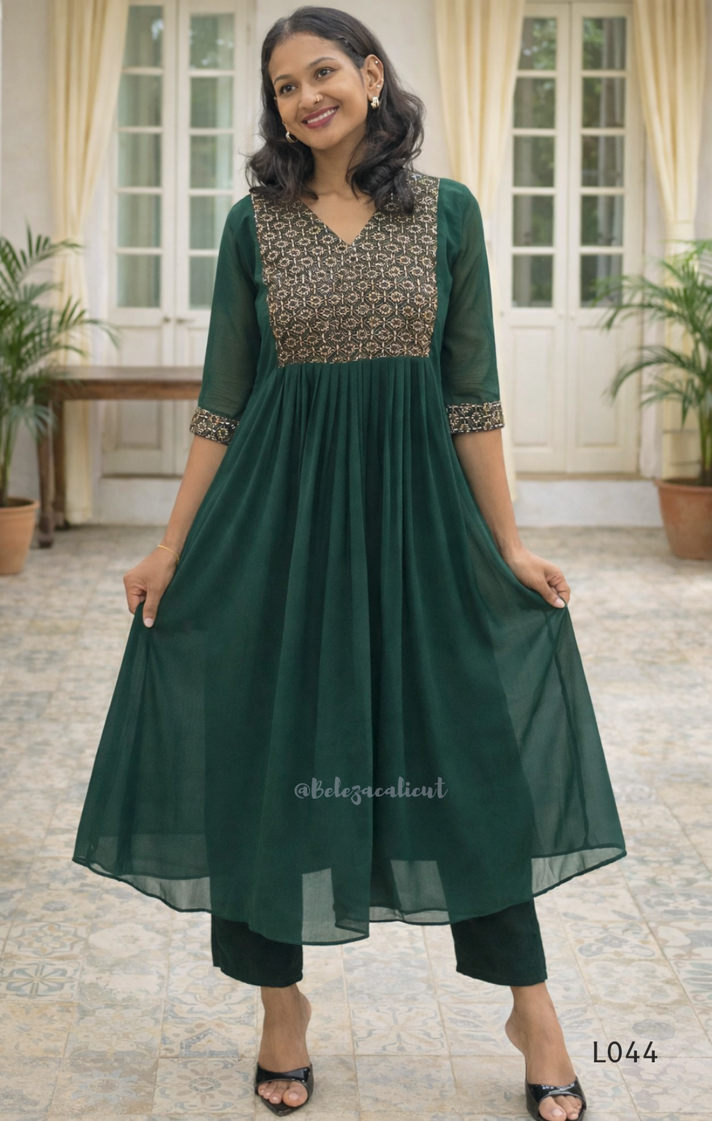 Woman wearing a green dress with a patterned top in a room with plants and a table.