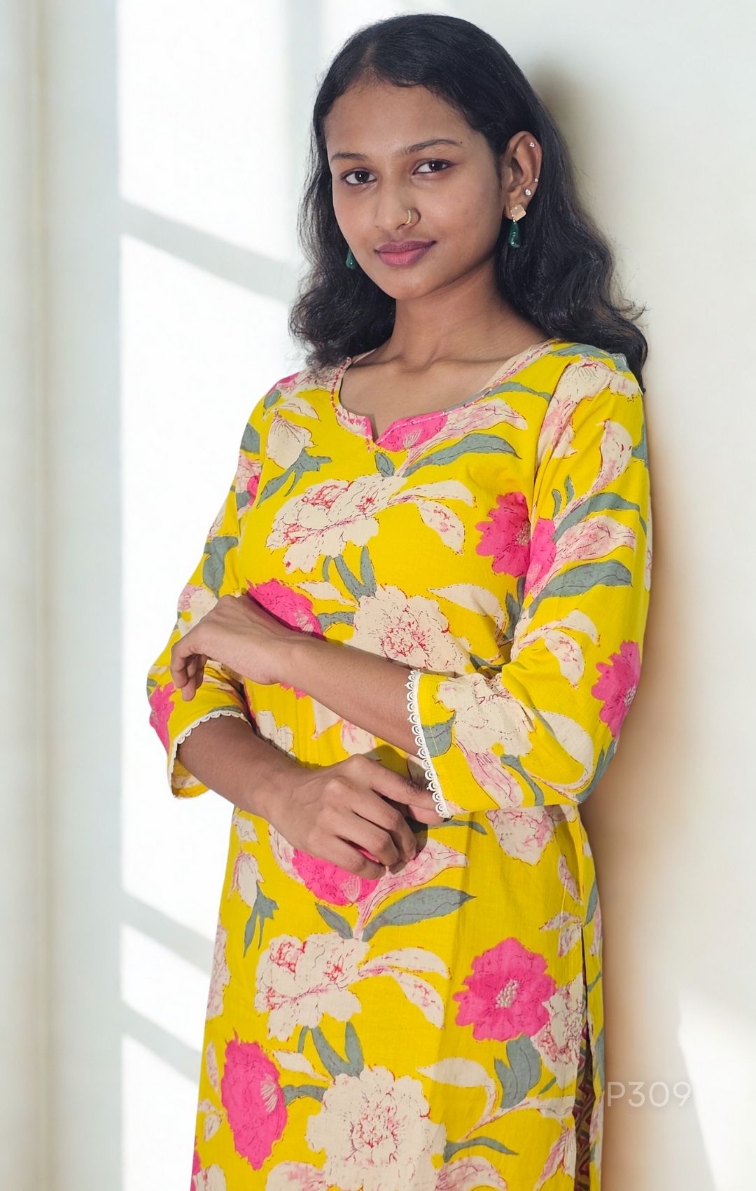 Woman wearing a yellow floral dress standing against a white wall.