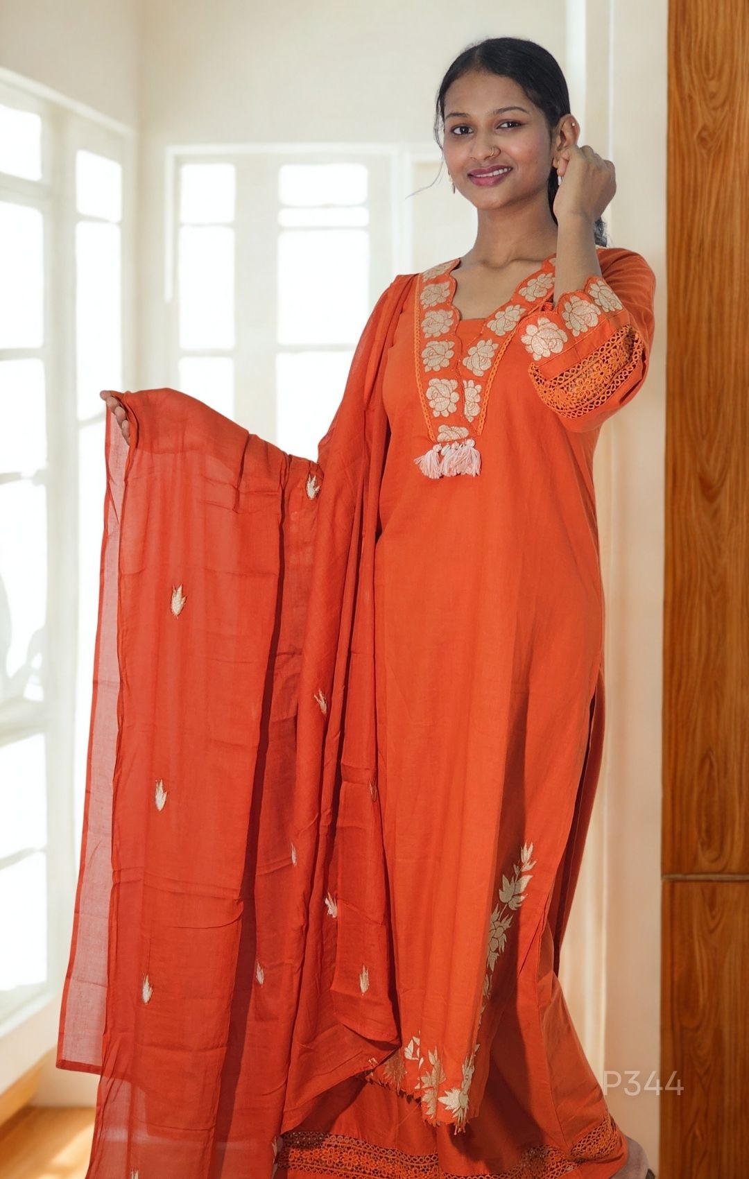 Woman wearing an orange traditional outfit with white embroidery in a bright room.