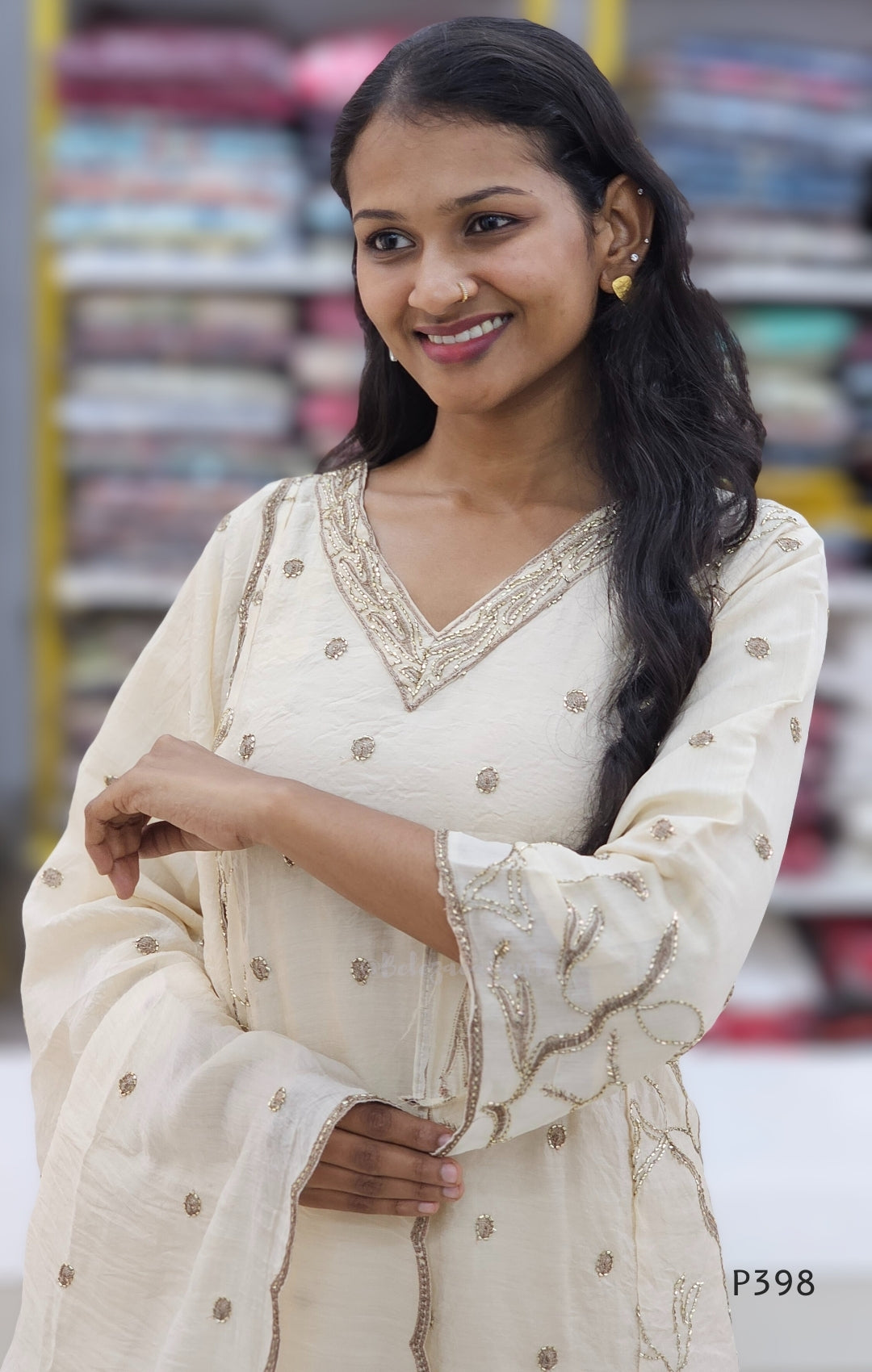 Woman wearing a cream-colored traditional outfit with intricate designs, standing in front of a blurred background.
