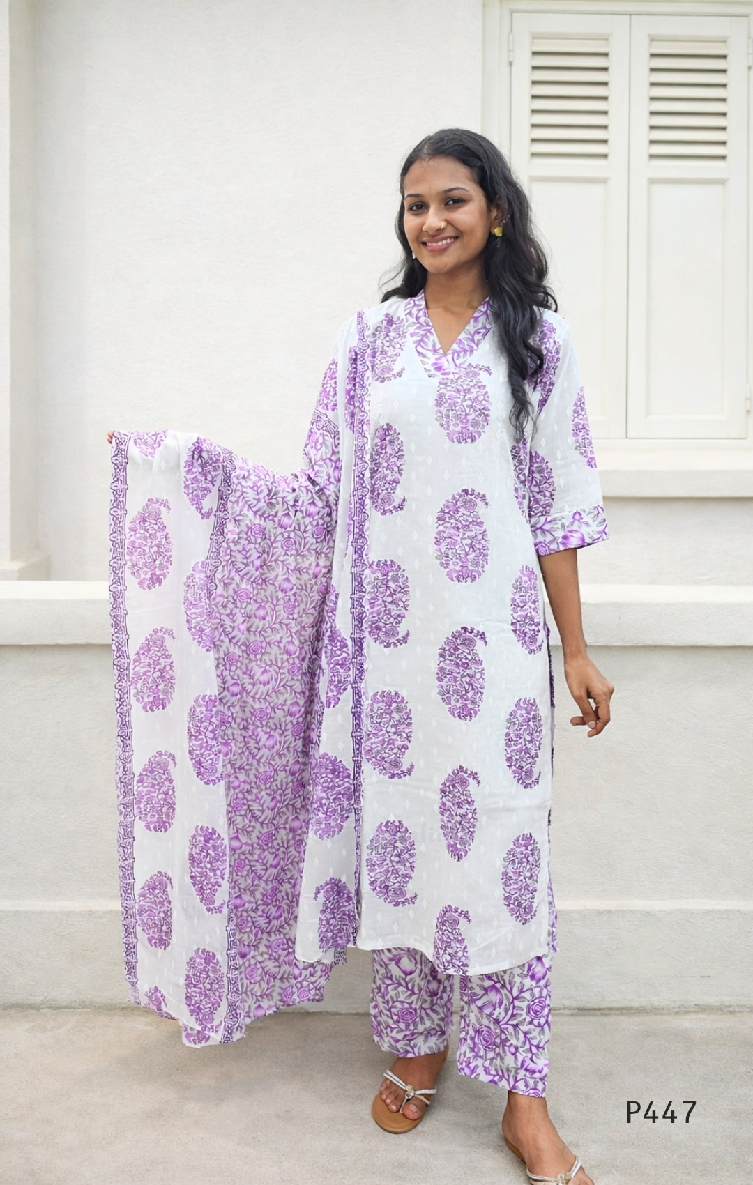 Woman wearing a white outfit with purple floral patterns in front of a white wall.