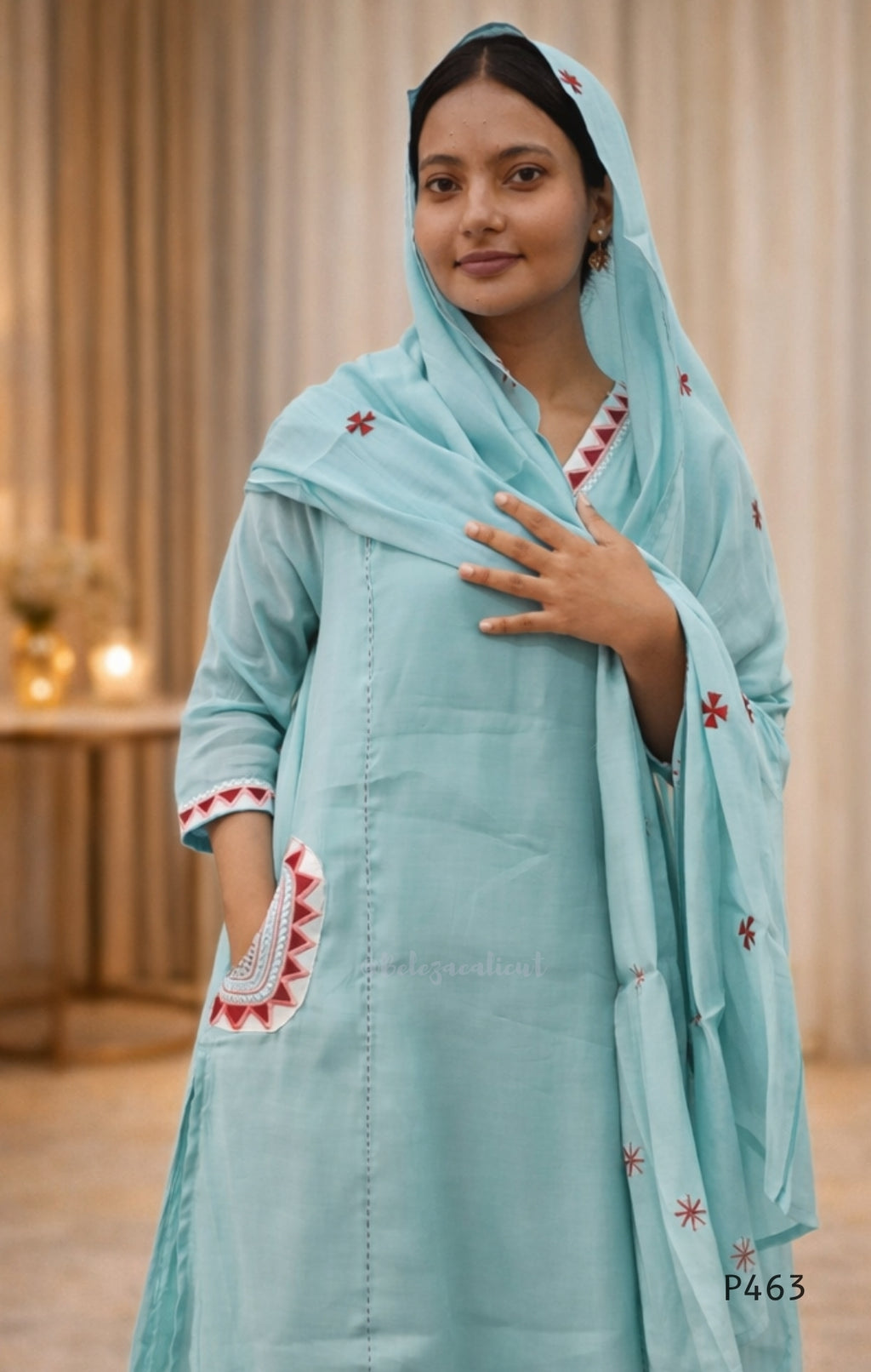 Woman wearing a light blue traditional outfit with red and white embroidery in a neutral setting.
