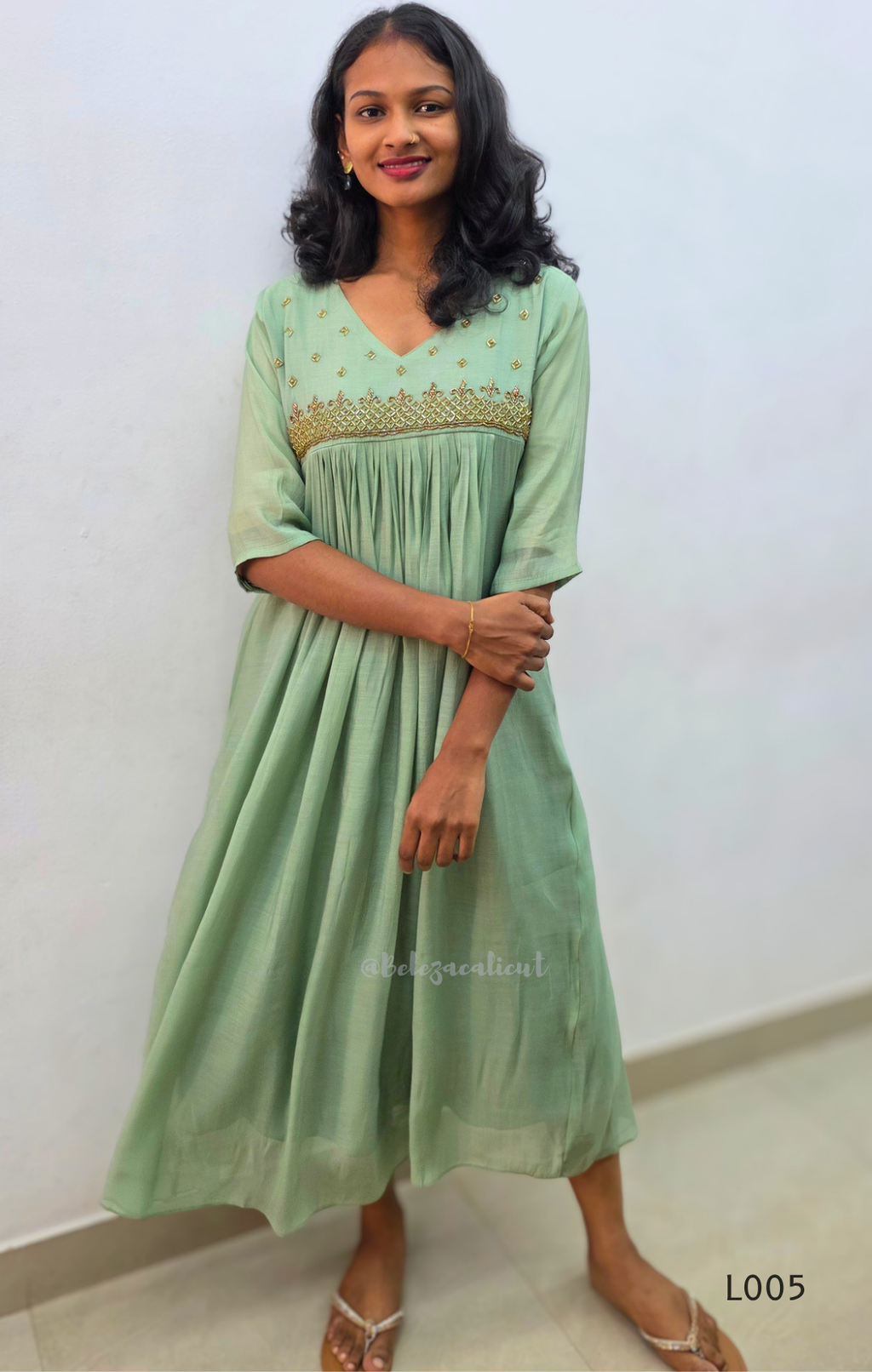 Woman wearing a light green dress with embroidery, standing against a white wall.