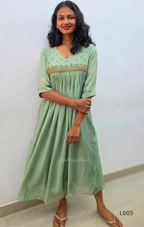 Woman wearing a light green dress with embroidery, standing against a white wall.