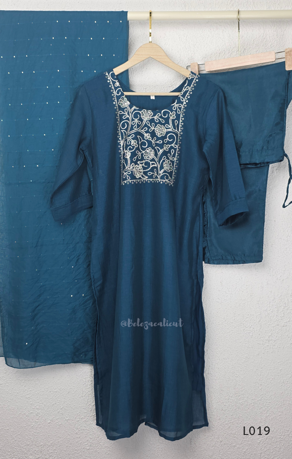 Deep teal blue silk blend 3-piece churidar set with square white floral threadwork embroidery yoke with bead droplet border, straight cut kurti with 3/4 sleeves, matching plain teal bottom with drawstring, and teal chiffon dupatta with scattered silver sequin dots, Indian ethnic wear from Beleza Calicut